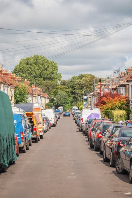A narrow, cobblestone street in Locksbottom lined with colorful, quaint houses, some painted in yellow and pastel shades, with small front gardens decorated with potted plants and flowers. The houses