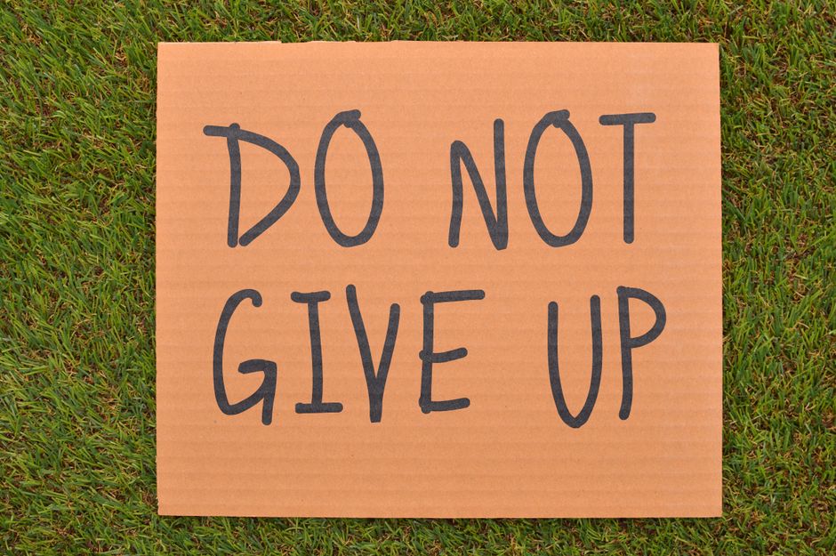 A rectangular cardboard sign with a light brown, corrugated surface displaying the handwritten message 'DO NOT GIVE UP' in large, dark marker letters. The sign is positioned on a grassy outdoor area, with green blades of grass visible around its edges. The setting appears to be in a residential or outdoor space, and the sign may symbolize encouragement or motivation, often used during home relocation or moving processes to promote a positive mindset. The natural lighting suggests daytime. This image aligns with home removals and furniture transport services provided by Man with Van Locksbottom, supporting themes of packing, moving, and loading during house removals.