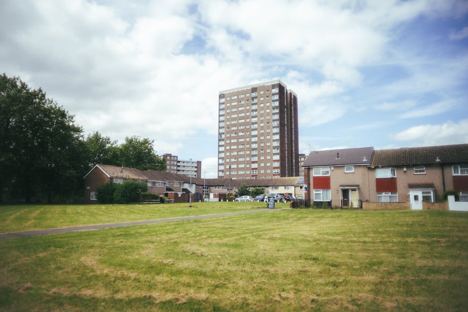 A wide view of a grassy area in a residential neighbourhood during daytime, with a row of small, single-story houses with brick walls and tiled roofs in the foreground. Behind the houses, a tall, multi-storey apartment building with numerous windows dominates the scene, set against a partly cloudy sky. A few cars are parked near the houses, and the open green space appears to be used for outdoor activities or as an area for loading and unloading furniture during a house removal. The setting is well-lit with natural daylight, and there are some trees on the left side of the image. This scene exemplifies the type of environment where a professional removals company such as Man with Van Locksbottom might assist with home relocation, furniture transport, or packing and moving services, especially within urban and suburban areas in Locksbottom.