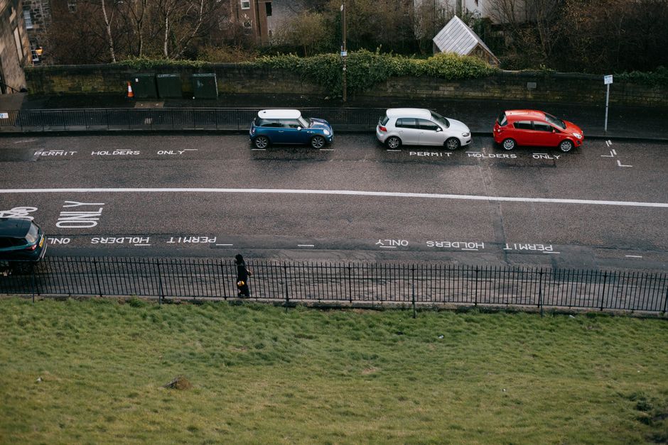 An aerial view of a street featuring three parked cars: a blue compact vehicle, a white hatchback, and a red hatchback, all aligned along a designated parking zone marked with 'Permit Holders Only' and 'Unload' signage painted on the asphalt. Behind the parked cars, there is a metal fence separating the street from a grassy area with a pedestrian walking along a sidewalk. The street appears to be in an urban residential area, with trees and shrubbery visible in the background. The scene is illuminated with natural daylight, capturing the typical environment where home relocation and furniture transport services by Man with Van Locksbottom might operate, including loading and unloading processes essential for effective removals.