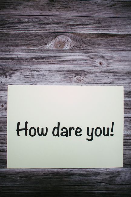 A rectangular sign with a white background and bold black text reading 'How dare you!' is placed on a weathered wooden surface with visible grain and knots. The wood has a natural, aged appearance with horizontal planks. The sign appears to be positioned flat on the surface, which may be part of a wooden table or outdoor decking. The lighting is natural, highlighting the texture of the wood and the contrast of the sign's lettering. This image could reference a humorous or confrontational message, often used in stationery or visual communication related to moving or household interactions, consistent with themes of relocation or house removals. The scene is simple, emphasizing the message on the sign against the rustic background, suitable for accessibility and SEO relevance to relocation services or moving-related content by Man with Van Locksbottom.