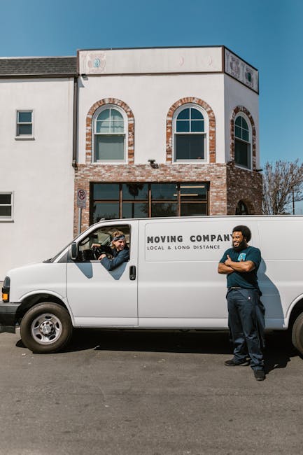 A white moving van parked on the street in front of a two-storey residential building, with two men from Man with Van Locksbottom engaged in a home relocation process. One man is inside the van, visib