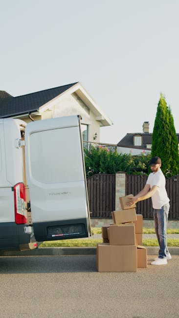 Two movers from Man with Van Locksbottom are engaged in the home relocation process, loading a large cardboard box onto a hand truck. The scene is set outdoors on a paved driveway, with the rear doors