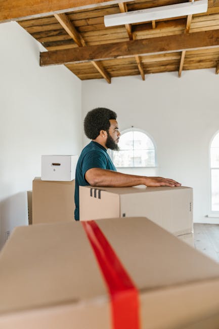 A man and a woman stand together indoors, smiling at the camera while holding a large cardboard box during a home relocation process. Behind them, there are several stacked cardboard boxes, some seale