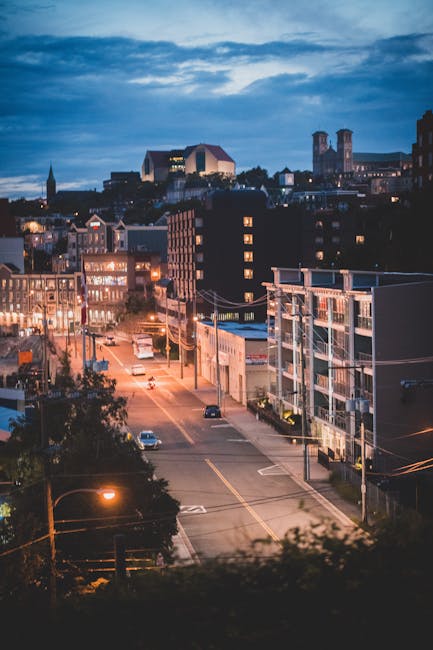 A twilight view of a suburban street in Locksbottom with residential and commercial buildings illuminated by streetlights and interior lighting. The scene features parked cars along both sides of the road, with visible facades of modern apartment blocks and retail units. In the background, there is a faint outline of historic landmarks and a church steeple against a partly cloudy sky. This urban setting depicts a quiet moment during early evening, with a sense of calm that aligns with home relocation activities such as moving or packing. The scene captures the typical environment where a professional removals company, like Man with Van Locksbottom, might organize furniture transport and packing services, highlighting the importance of planning in house removals in BR6.
